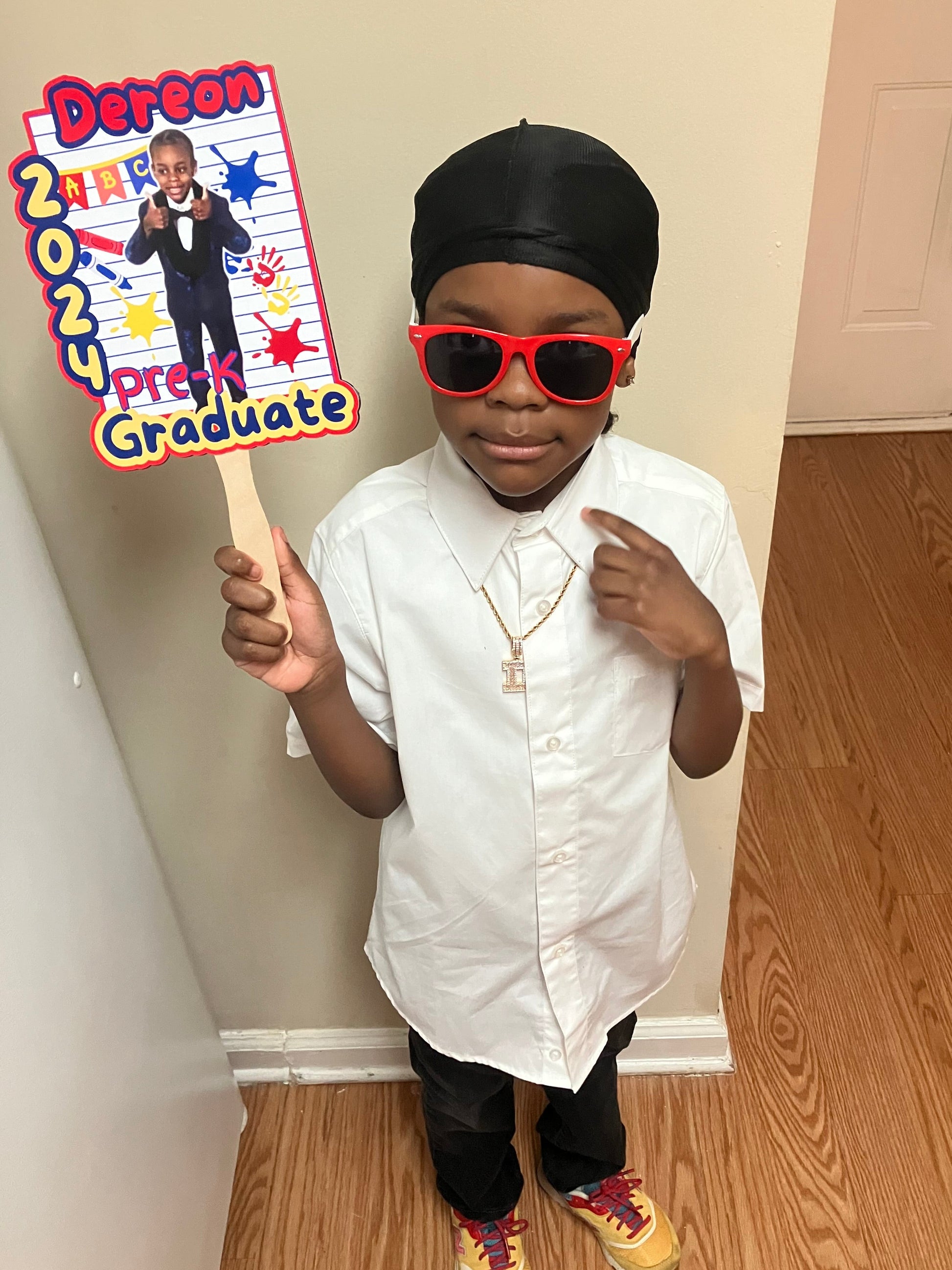 Person in a white graduation gown holding a '2024 Graduate' sign indoors.